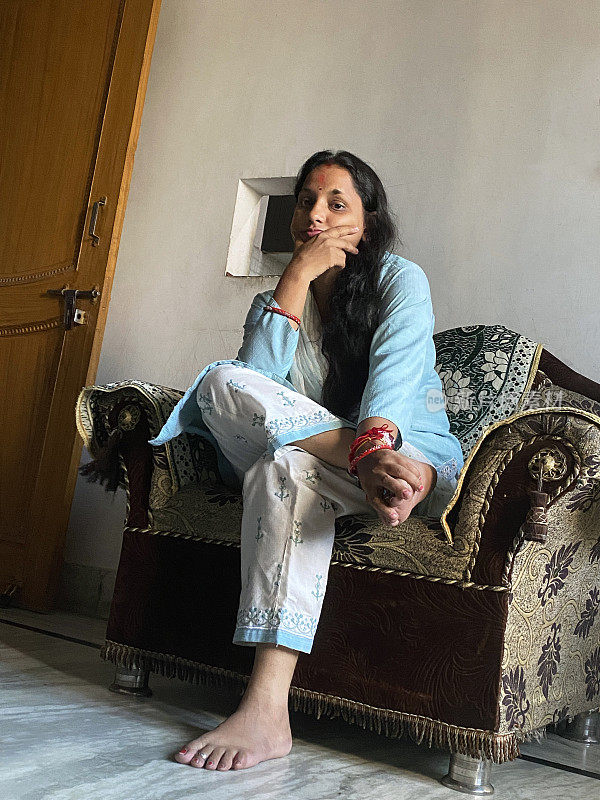 穿着Salwar Kameez传统服装的印度妇女的形象，坐在休息室的扶手椅上，一条腿交叉在膝盖上，看着相机，专注于前景